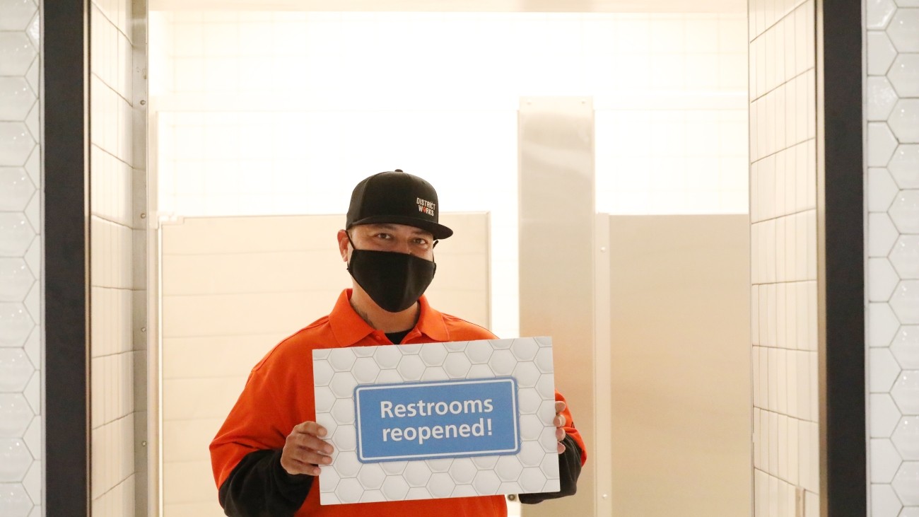 District Works worker holds sign saying "Restrooms Reopened!" in front of the Powell BART Station restroom.