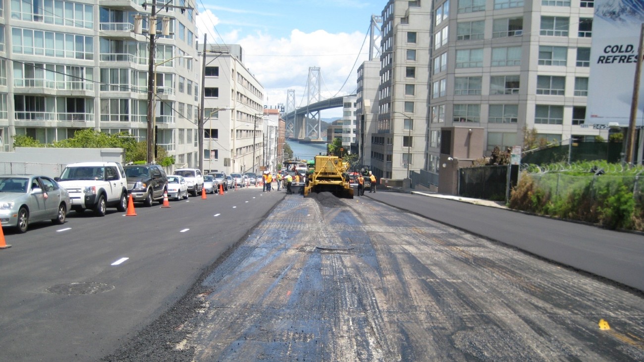A street is being repaved, with workers using heavy equipment to work on the center lane. The Bay Bridge is visible in the background.