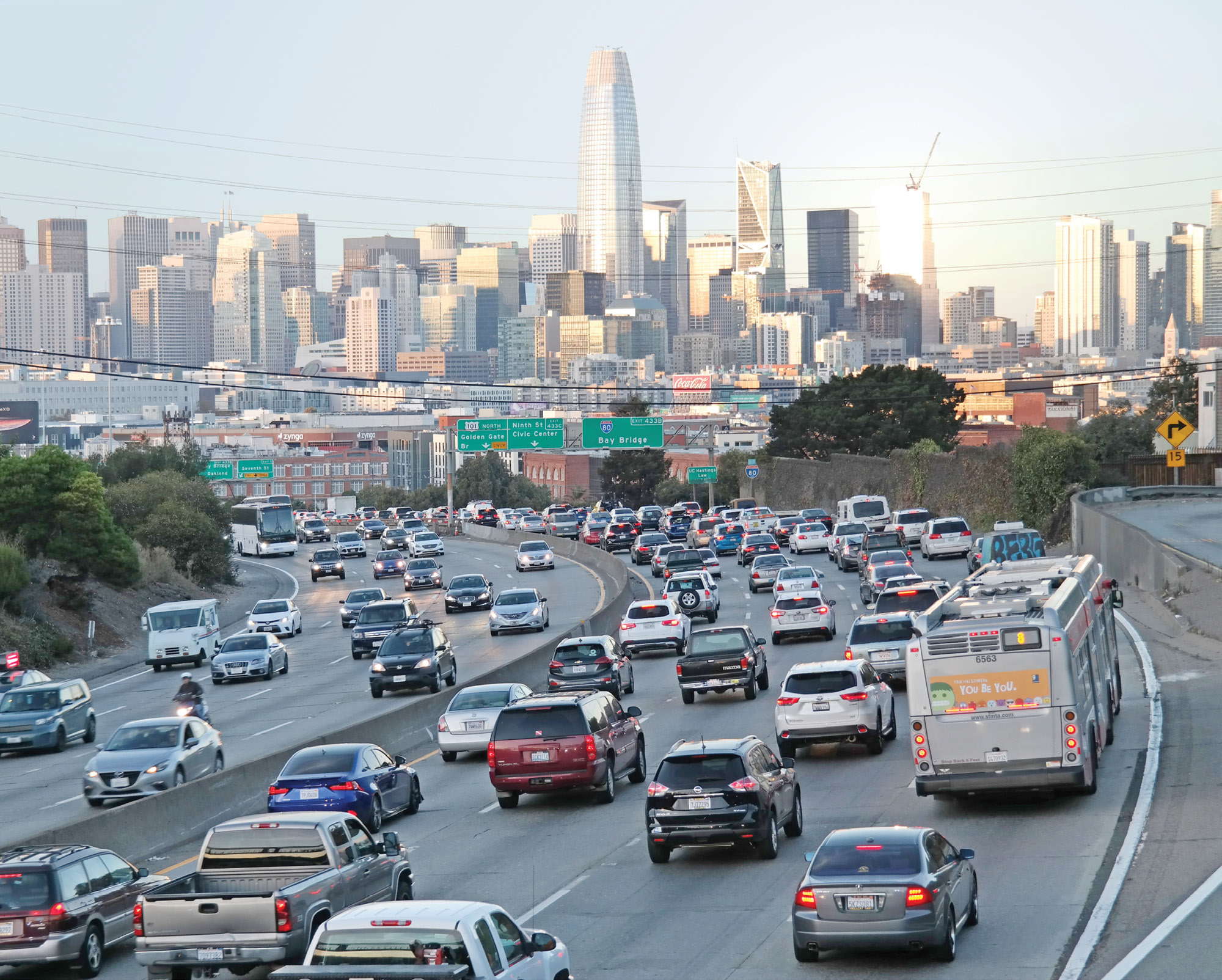 A view of I-80 filled with cars, trucks, and a Muni bus, seen from the 18th Street pedestrian bridge. The San Francisco skyline is visibe in the background.