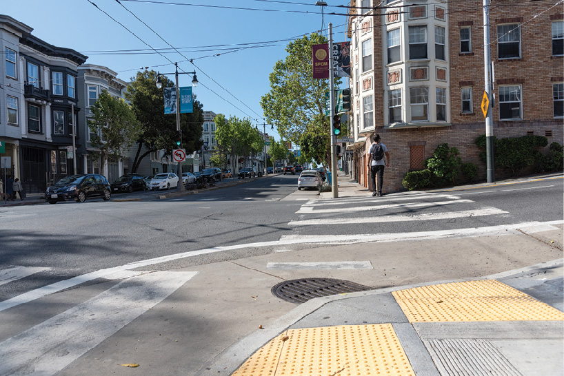 A close up of a street corner. The sidewalk has yellow tactile strips. There is a person crossing the street at a crosswalk. There is a beige painted area wrapping around the street corner.