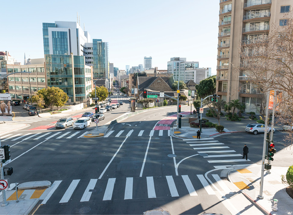An overhead view of an intersection in Cathedral Hill. There are multiple lanes of traffic in each direction. There are newly painted white crosswalks across all of the streets. Two pedestrians are crossing the street on the right side of the photo. The background has some tall apartment and office buildings.