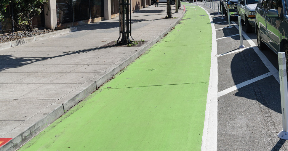 A green-painted bike lane is separated from traffic by a painted buffer, flex-hit posts, and parked cars.