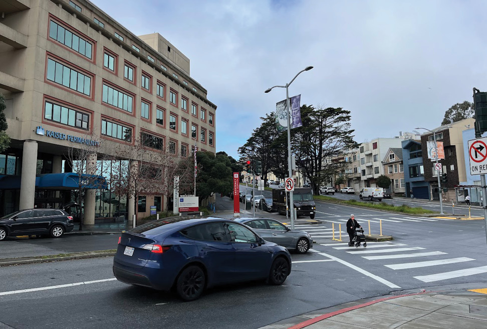 A person using a walker uses the crosswalk to cross a wide street. There is a Kaiser Permanente medical facility in the background on the left side of the photo. In the foreground, there are two cars waiting at a red light.