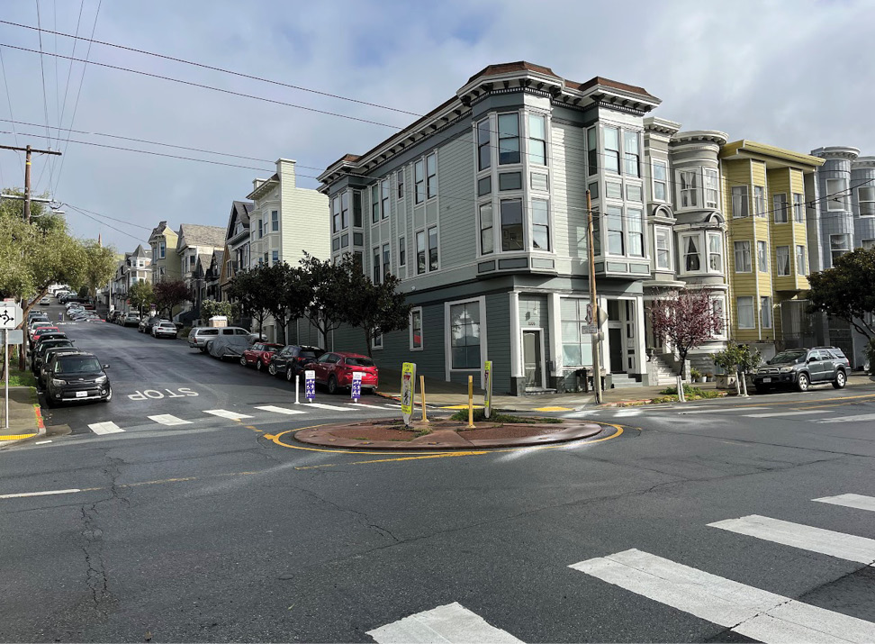 A roundabout, paved with red brick, in the middle of an intersection. The background has a three-story apartment building. One of the streets leading away from the roundabout is a slow street, with purple signs labeled “Slow Street” in the middle of the street.