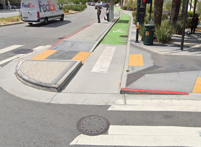 Close up of a concrete island separating a bike lane from the street. The entrance to the bike lane is a sharp, 90-degree corner. There is yellow tactile paving on either side of the concrete island and leading onto the sidewalk. The bike lane is painted green in the background.