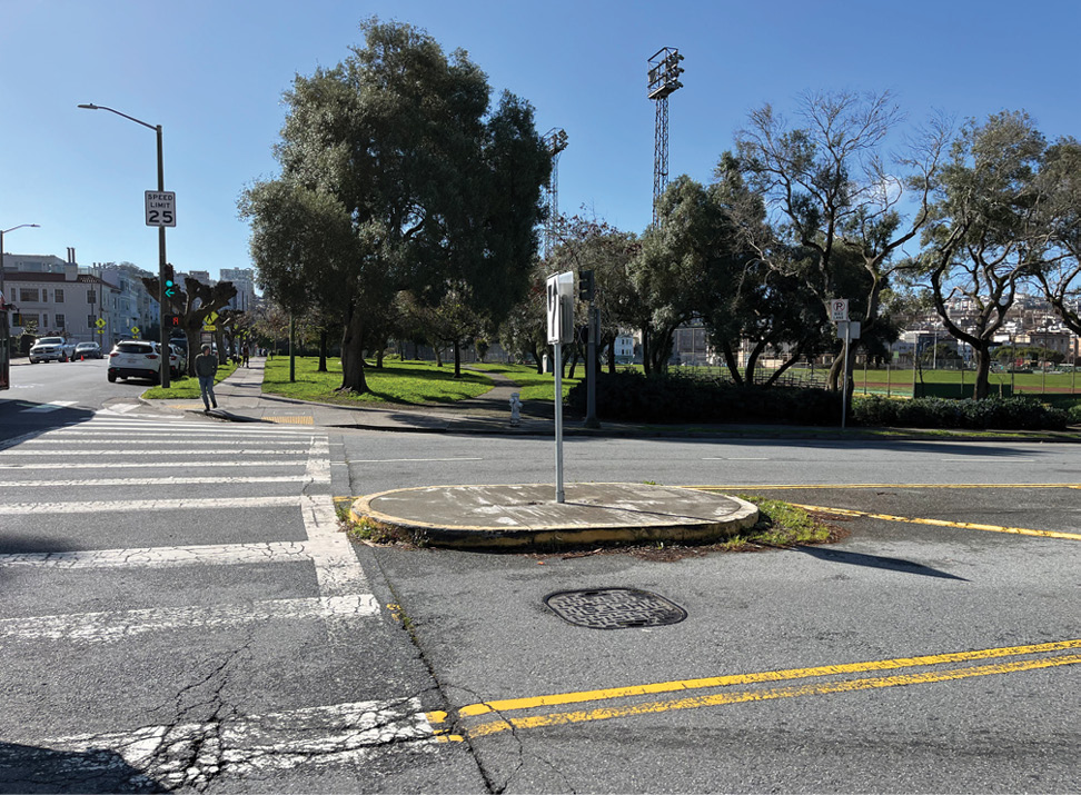 A concrete traffic calming median in the middle of a wide street. There is a crosswalk on the left side of the photo and a park in the background.