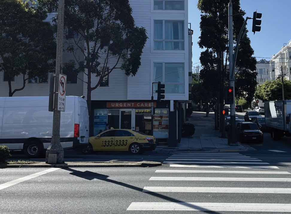 A crosswalk across a wide street. There is a concrete traffic calming median on the left side of the photo. The background shows an urgent care medical facility with a taxi and van parked outside.
