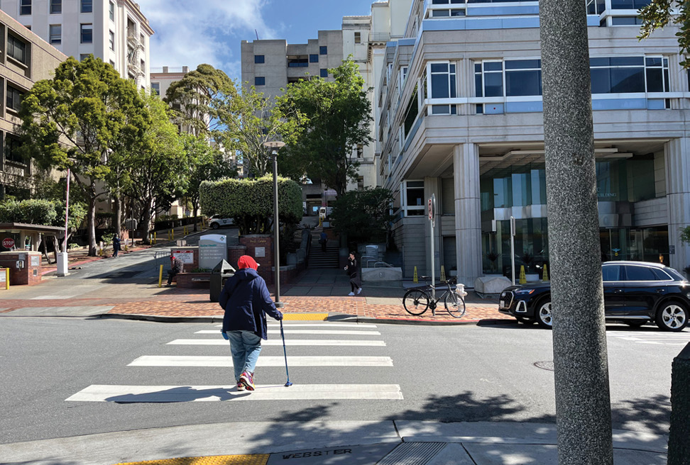 A person using a cane crosses the street at a crosswalk. The crosswalk leads to a medical facility in the background. There is a bike parked in front of the medical facility and a car parked in the street.