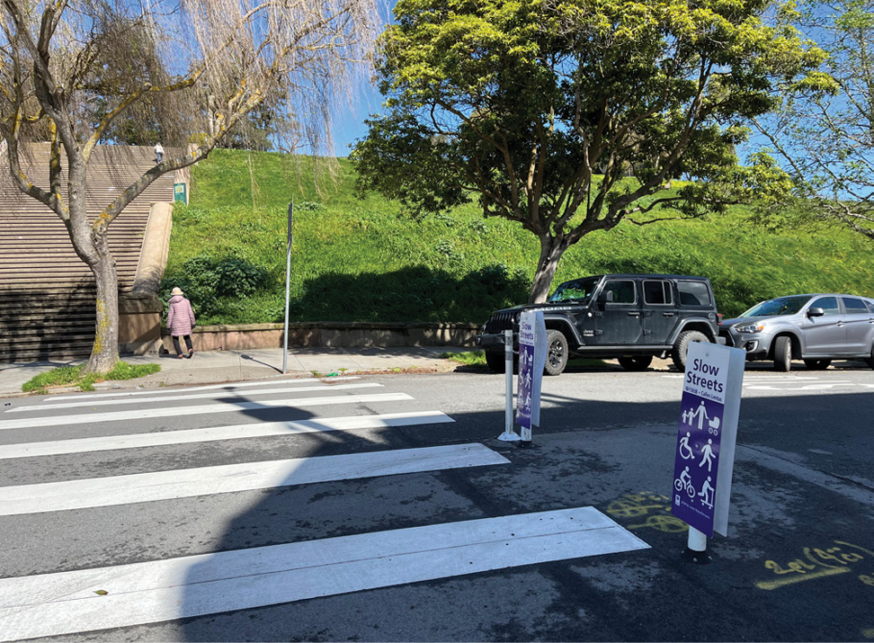 A crosswalk leading to the entrance of a park in the background of the photo. There is an older woman with a walker on the sidewalk in front of the park. There are two signs next to the crosswalk in the street that say “Slow Streets” with icons of people walking, biking, and rolling.