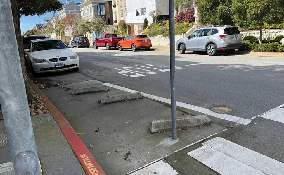 A set of three small concrete curbs blocking off a parking space at the corner of an intersection. The sidewalk curb is painted red with a yellow SFMTA logo. There are a number of cars parked in the street in the background.