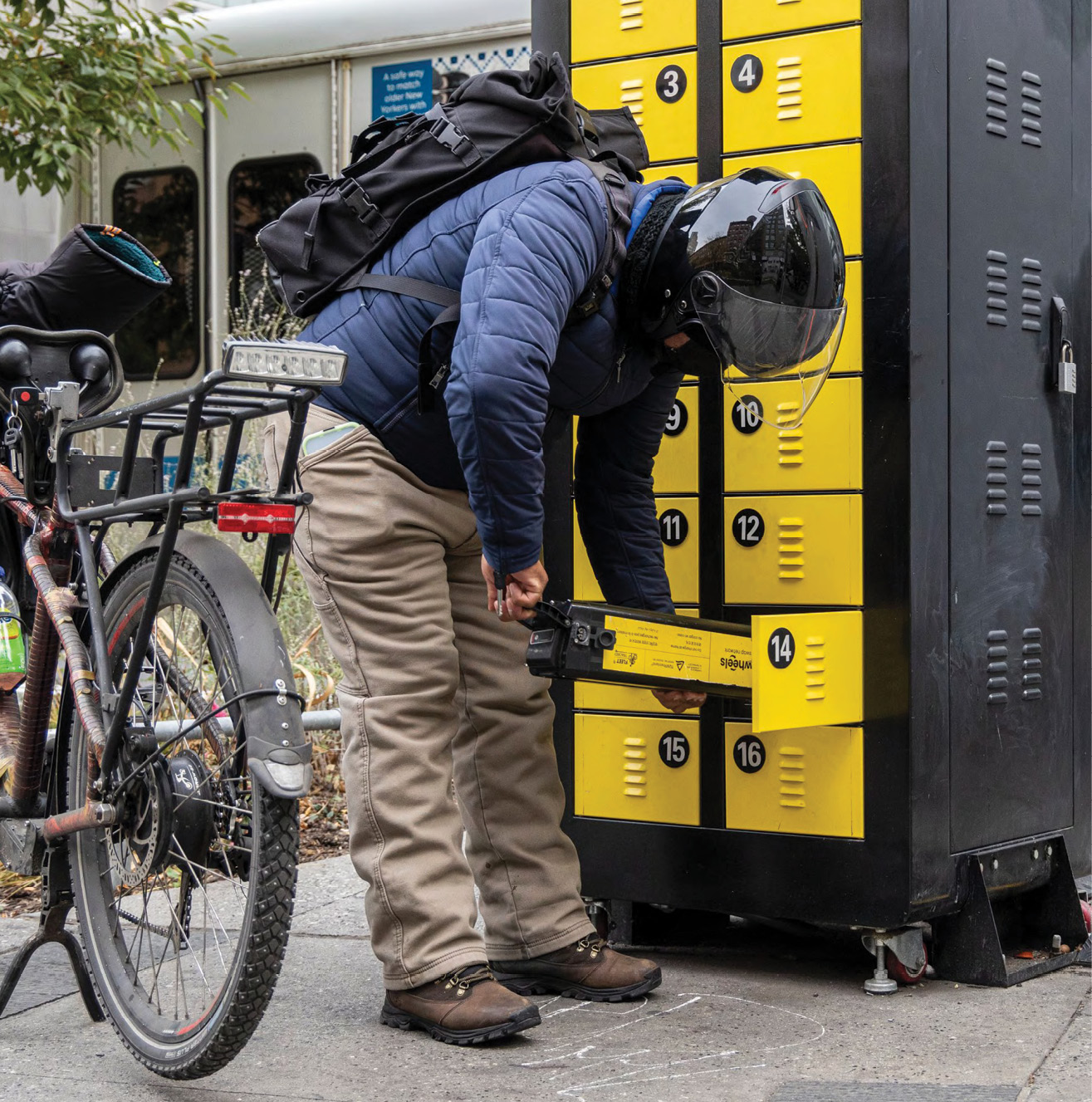 An ebike deliver worker bending over to retreive a battery from a yellow locker. The workers' bike is propped up next to them.