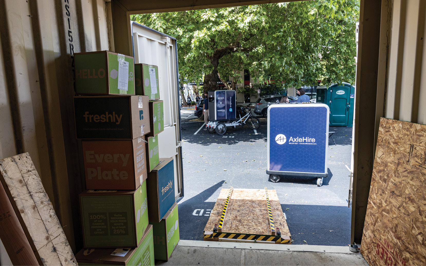 A view from the inside of a metal storage container with boxes stacked inside and a ramp leading to the exterior parking lot. A cargo tricycle is visible across the parking lot.