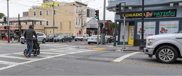 A man pushes a stroller in a crosswalk. A truck waits to cross in the other direction at a limit line that has been painted more than ten feet before the crosswalk.