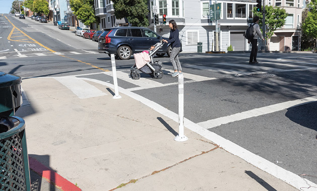 A woman pushes a baby stroller in a crosswalk. An area around the curb, approximately six feet wide, is painted tan with plastic soft-hit posts along its edge.