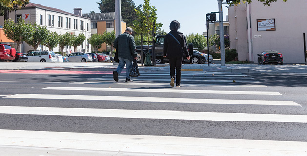 Two people walk in a crosswalk, which is painted with many wite stripes running perpendicular to the crossing direction.