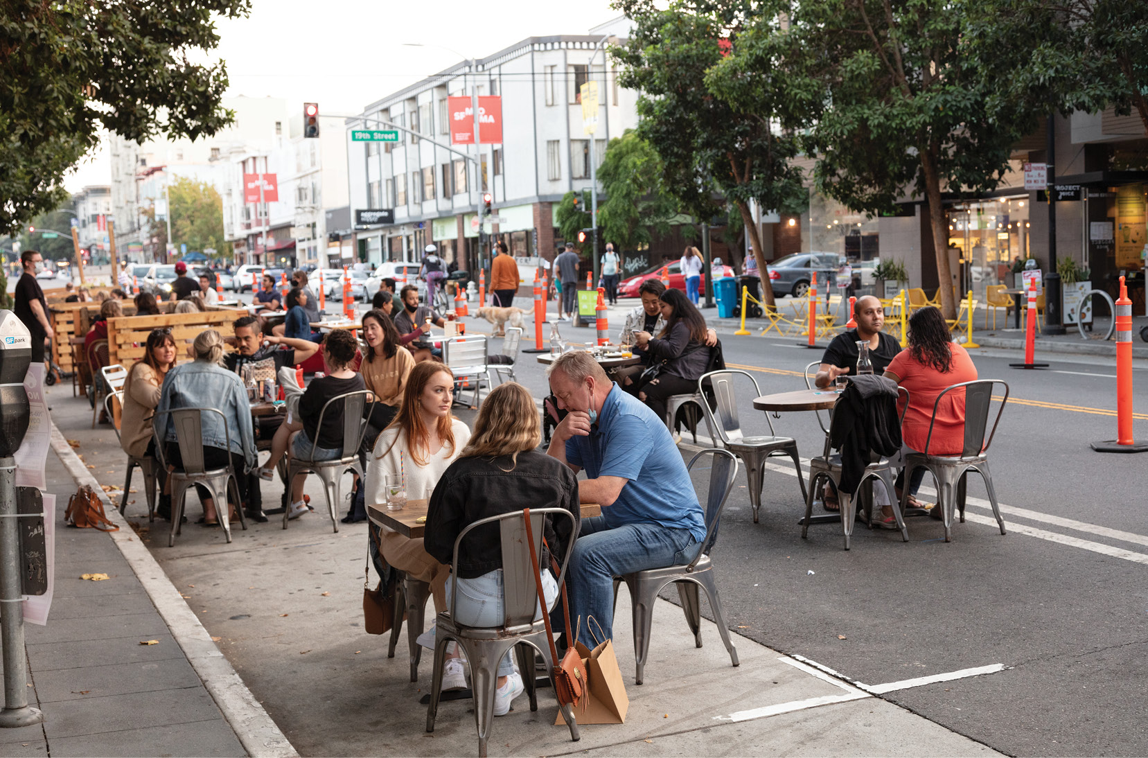 Diners sit at temporary tables set up in the street. There are small groups of two to five people seated at each set of tables. There are orange cones in the background that prevent cars from driving on the street.