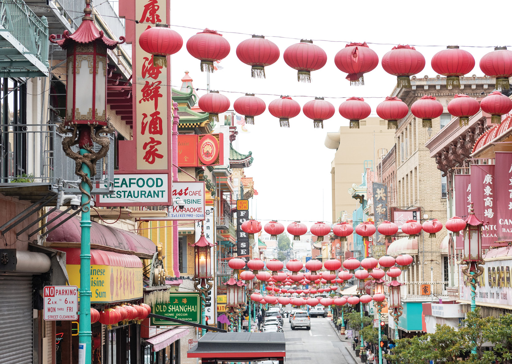 View looking down Grant Avenue in Chinatown. In the foreground, there is a street lamp that is decorated with a Chinese-style lantern and dragons around the lamppost. There are strings of red lanterns hanging horizontally across the street, all the way up and down the street.