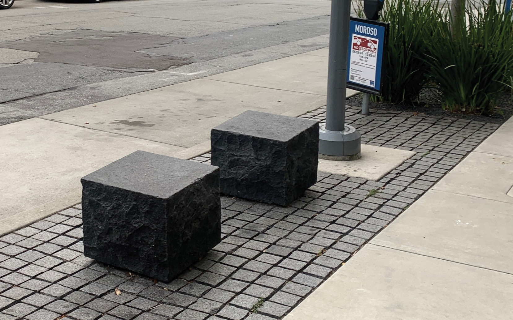 A close up of two small seats on a sidewalk. The seats are small stone cubes. They are mounted on the edge of the sidewalk on top of a strip that has been paved with small stone tiles. The sidewalk on either side is beige.