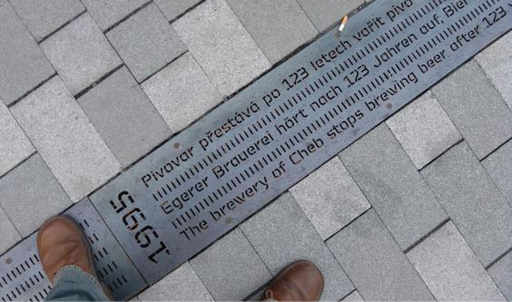 A top-down view of a historical marker inlaid on a sidewalk and a person’s feet standing on the sidewalk. The historical marker is a metal drain with the year 1995 inscribed and text in German. The sidewalk is paved with light grey tiles.