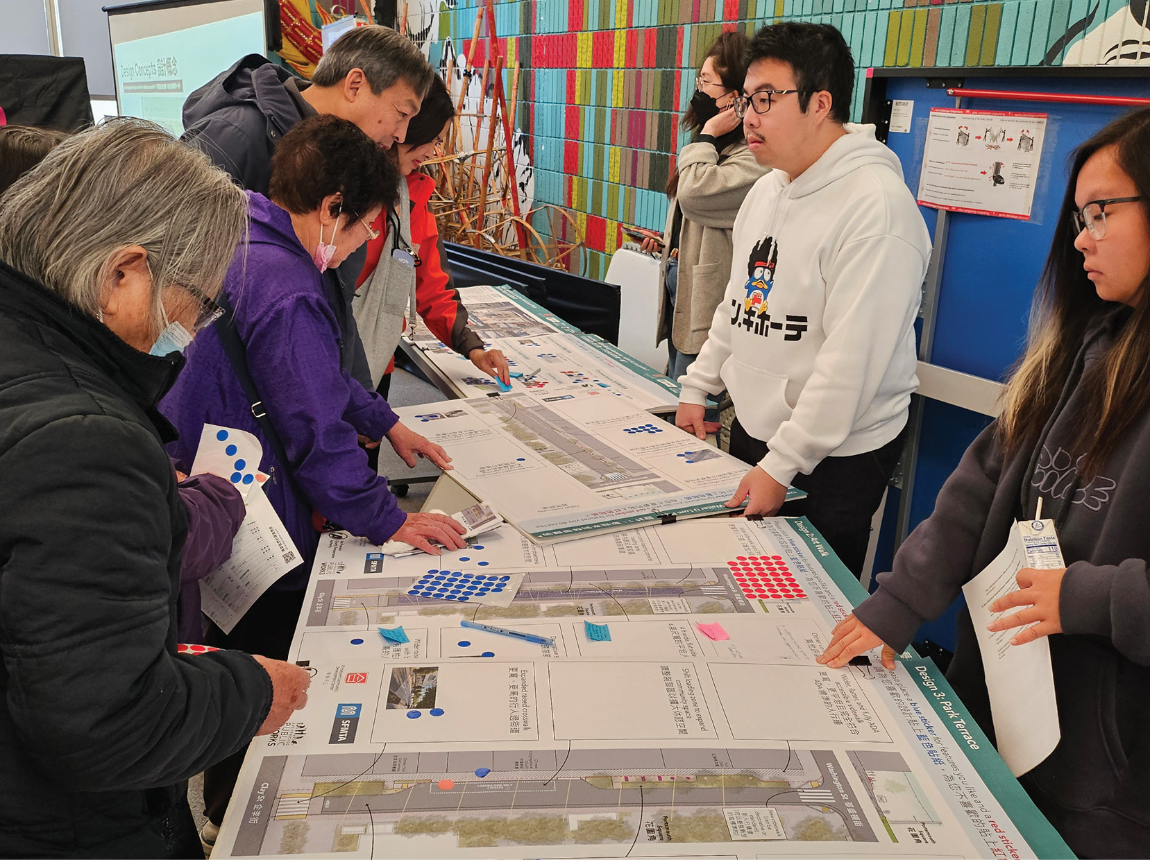 A series of boards depicting separate proposals for the Walter U Lum Alleyway are on the table. A number of members of the public are looking at the boards around the table. Some of the individuals have red and blue circle stickers and are sticking them on elements they do/don’t like. They are in a space with colorful red and blue tiles behind them.