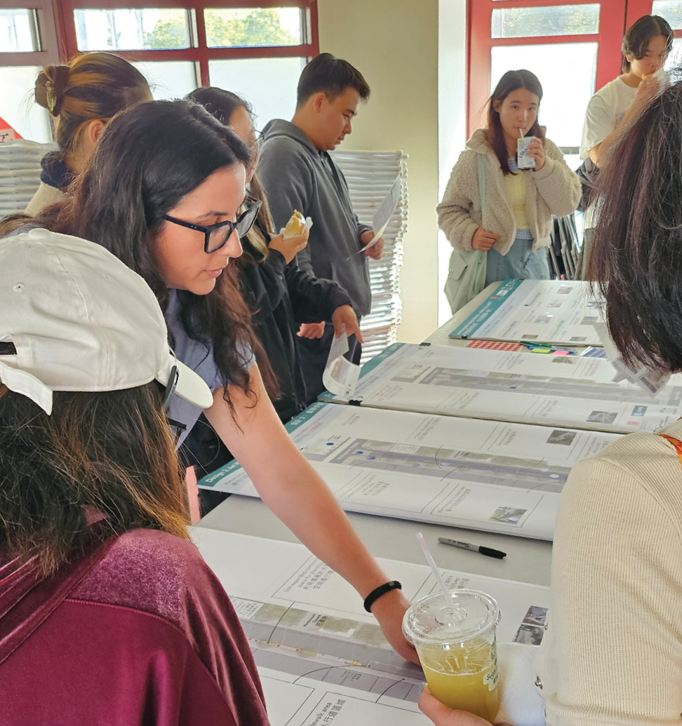 A series of poster boards depicting separate proposals for the Walter U Lum Alleyway are on a plastic folding table. A number of members of the public as well as some staff members are gathered around the table looking at the boards and paper handouts with a summary of designs printed on them. They are in a community room with windows behind them.