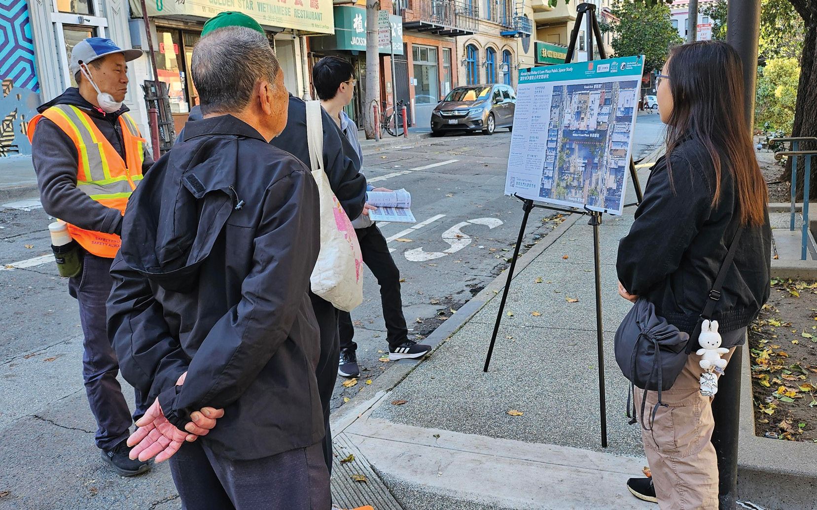 Two youth and a number of older men in jackets look at a poster board with a map of Walter U Lum alleyway on an easel. They are standing outside at an alleyway.