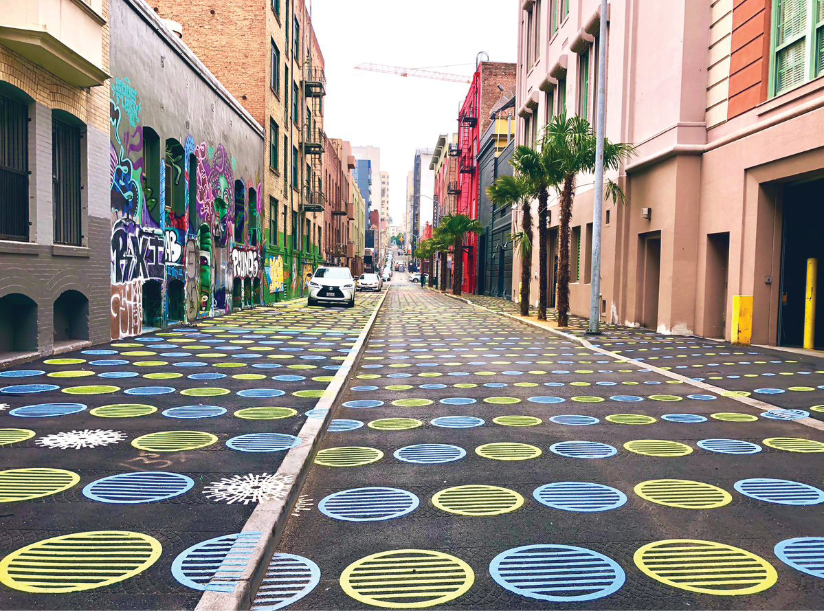 An image of Fern Street, an alleyway in San Francisco. The ground of the alleyway is decorated with a pattern of green and blue circles painted onto the asphalt. There are also trees lining the alleyway and a row of parked cars on the left.