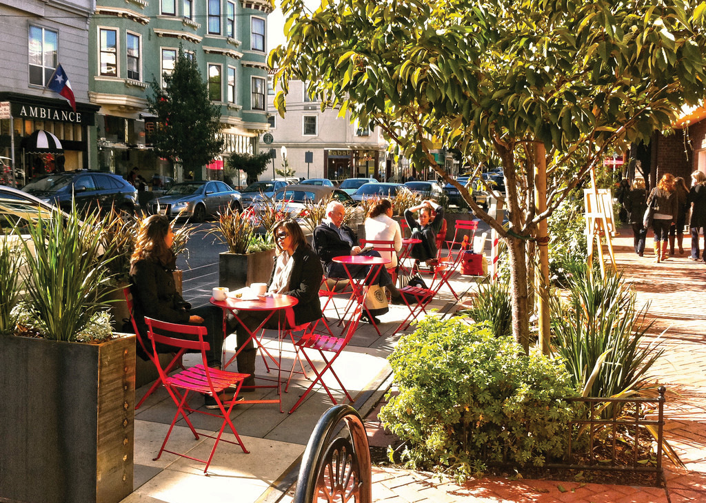 A parklet in San Francisco. There are red tables and chairs set up on an extension of the sidewalk. The parklet is separated from cars in the street by wooden planters. There are trees separating the parklet from the rest of the sidewalk, where there are a few pedestrians.