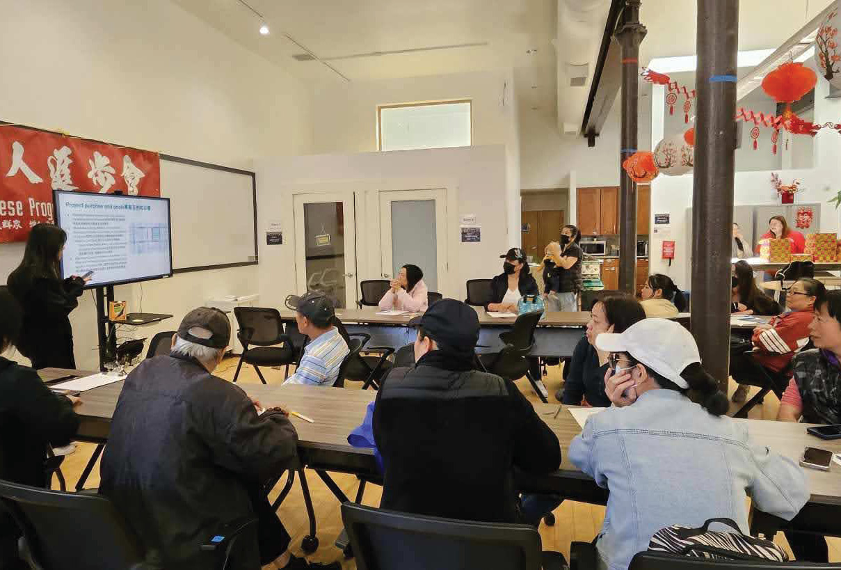 A community meeting with SRO Families. Members are seated around tables in a classroom-like setting. Two presenters stand in front of a screen, discussing a project purpose and goals, with text displayed in both English and Chinese. The room is decorated with red paper lanterns and traditional ornaments. Attendees, some wearing face masks, listen attentively and take notes.