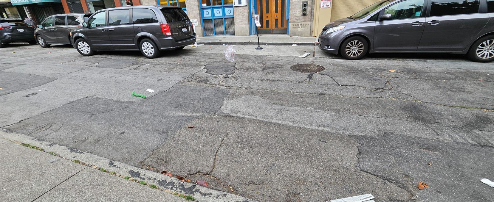 A view of the street surface on Walter U Lum Place. The pavement is cracked in many places and littered with leaves, paper, and plastic bags. There are four cars parked in the loading zone in the background.