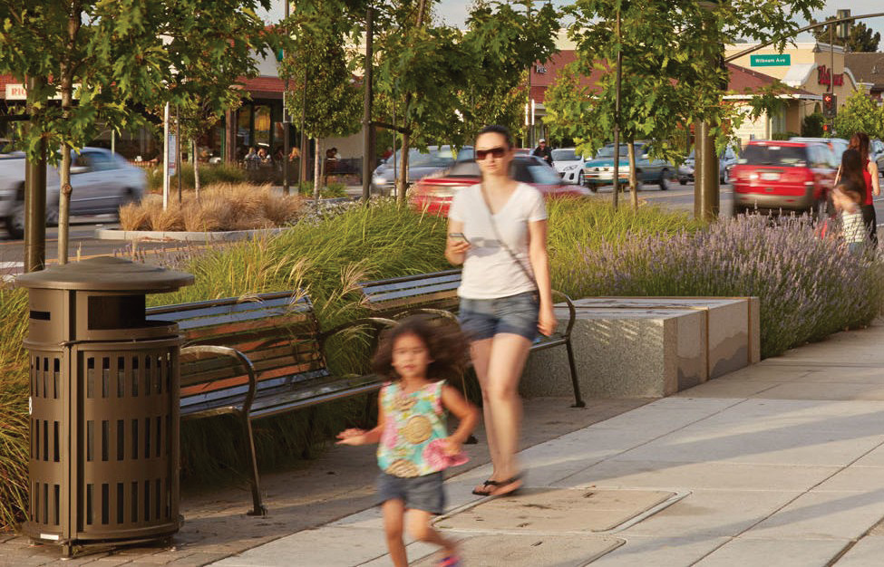 A view of a seating area next to a sidewalk. There is a young child running in the foreground and an adult in the background. The sidewalk is paved with concrete. There are two benches in the background in front of a planting area with long grass and trees.