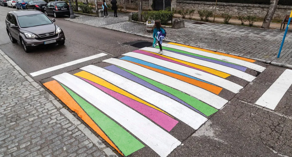 A close up of a decorative pedestrian crosswalk. There are colorful blocks painted in between the standard white markings of a crosswalk. There is a pedestrian crossing in the crosswalk and a sedan stopped in front of the crosswalk.