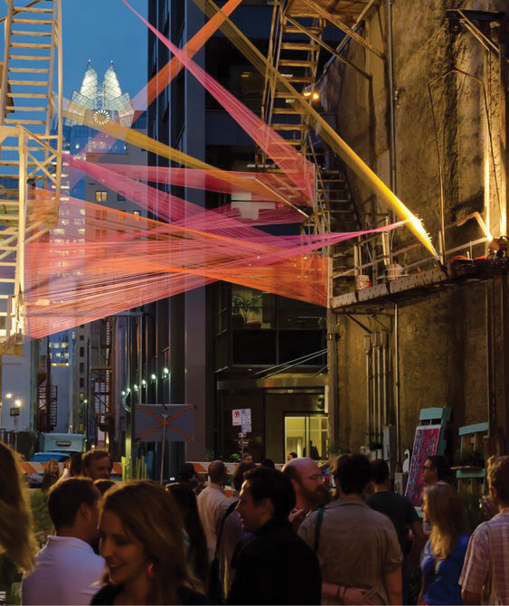A view of a crowded street at nighttime. There are many people standing in the street. There is an art installation of orange, yellow, and pink see-through fabric sheets criss-crossing the street, mounted to the sides of the building on either side.