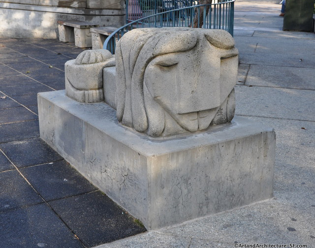 A close up of a stone sculpture in Portsmouth Square. The sculpture is a cube shape with an abstract face carved into the front. The sculpture is mounted on top of a larger stone base.