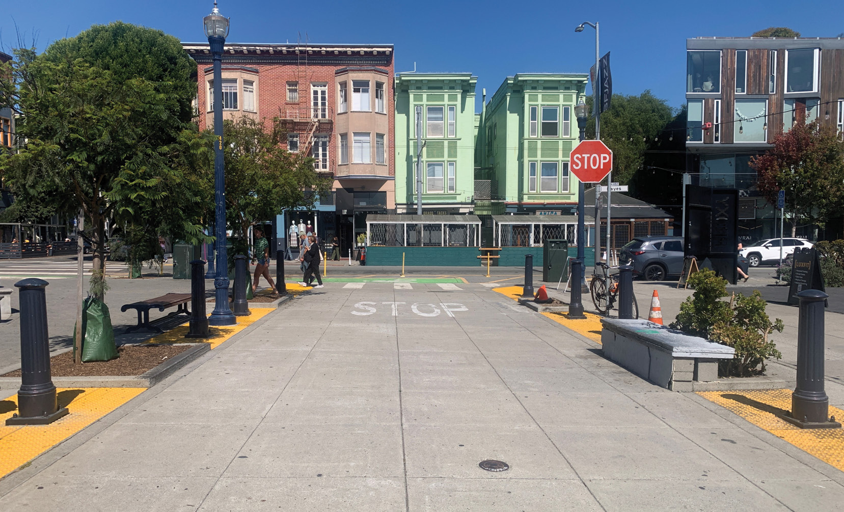 A view looking down a raised crosswalk at Patricia’s Green in Hayes Valley. The crosswalk is paved with concrete and there are yellow tactile strips on either side of the crosswalk, green bollards, stone benches, and trees. At the end of the crosswalk, there is a stop sign.