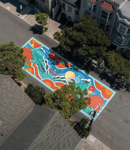 A top-down view of a colorful mural painted onto a street in San Francisco. The mural shows ocean waves in blue and teal, and there are orange poppies, the Golden Gate Bridge in the background, and a rising full moon. There are trees lining either side of the street.