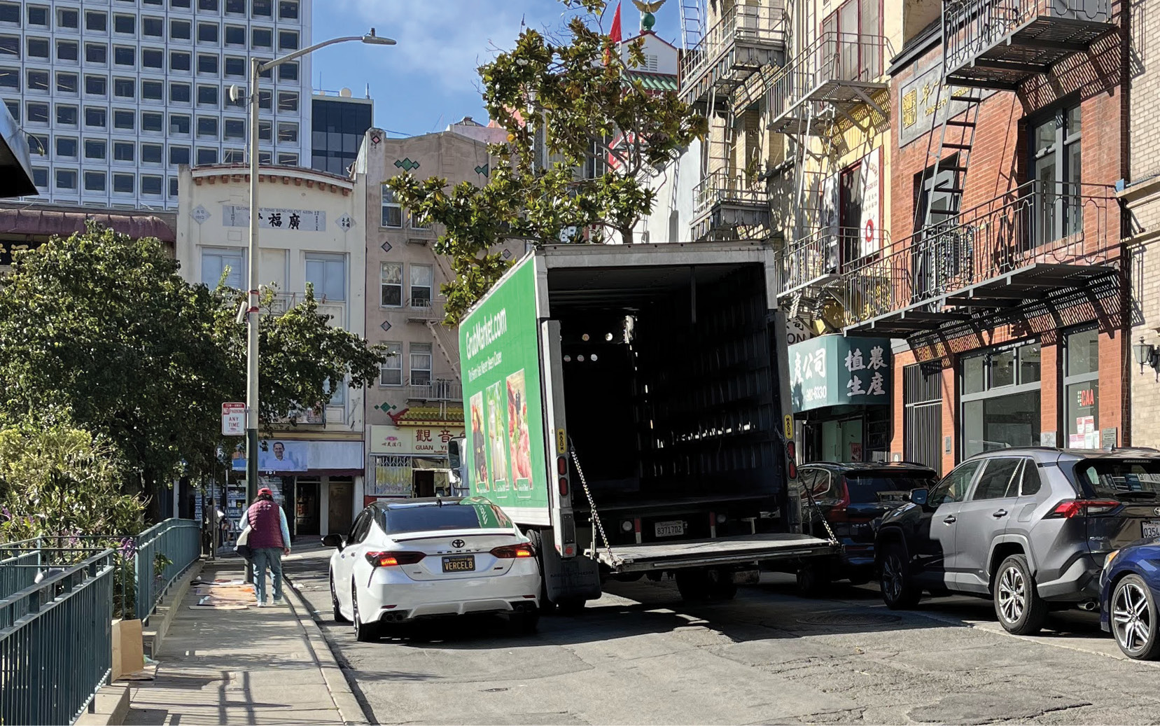 A view of Walter U Lum Place showing a full view of the street. There is a large, green box truck double parked in the street with the cargo area open. The loading zone to the right of the box truck is fully occupied by parked vehicles. A white sedan passes the box truck on the left side, using the remaining open space in the street. There is a person walking on the sidewalk to the left of the sedan.