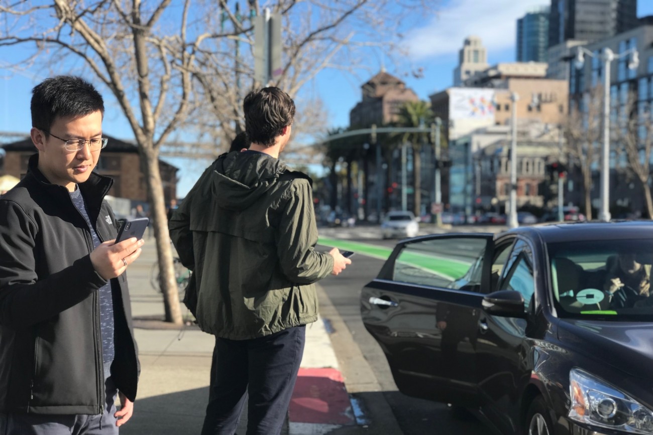 People standing on a sidewalk with phones and a ride-hail vehicle dropping someone off