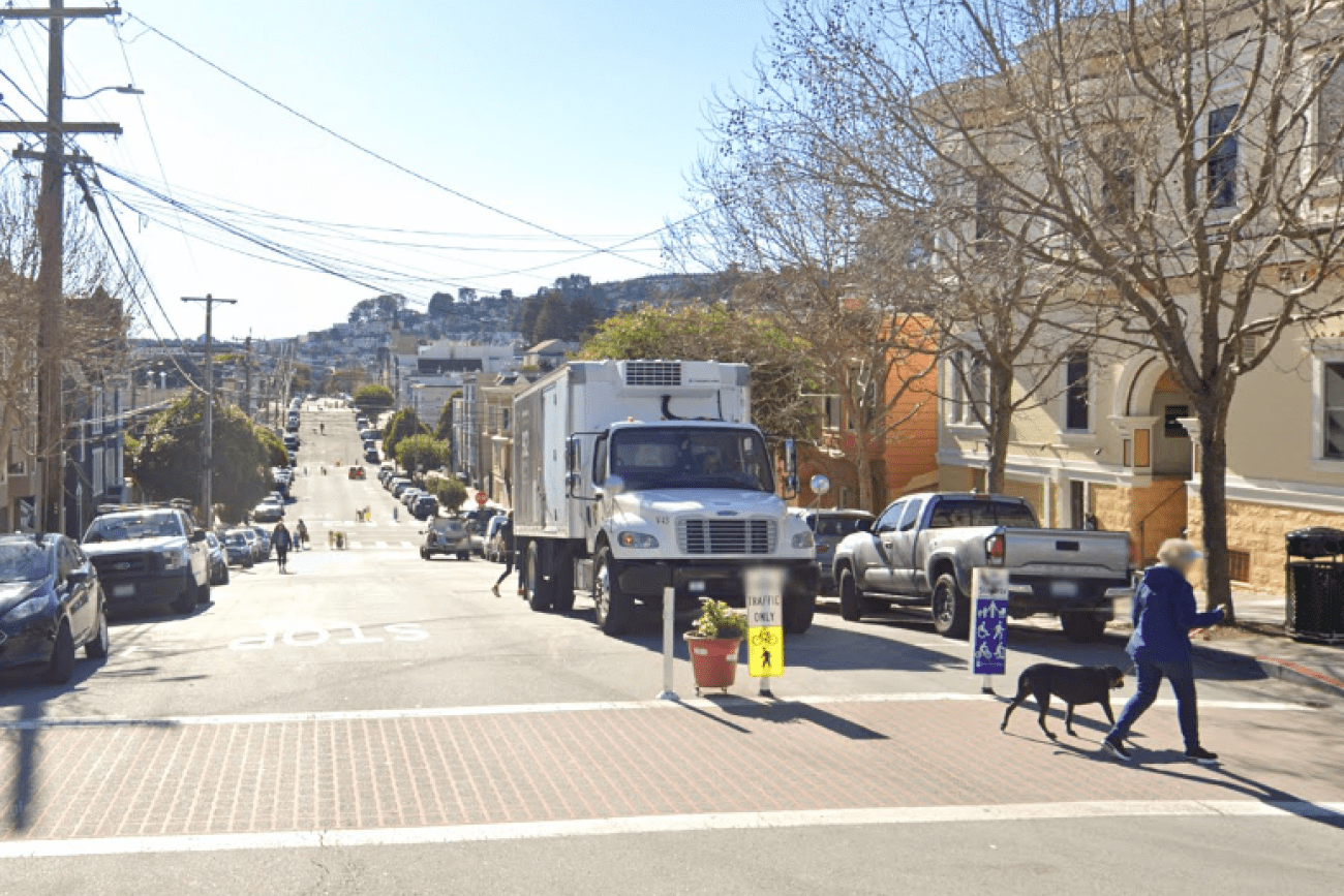 Street-level view of Sanchez Slow Street on a sunny day. A person walks a dog across the intersection.