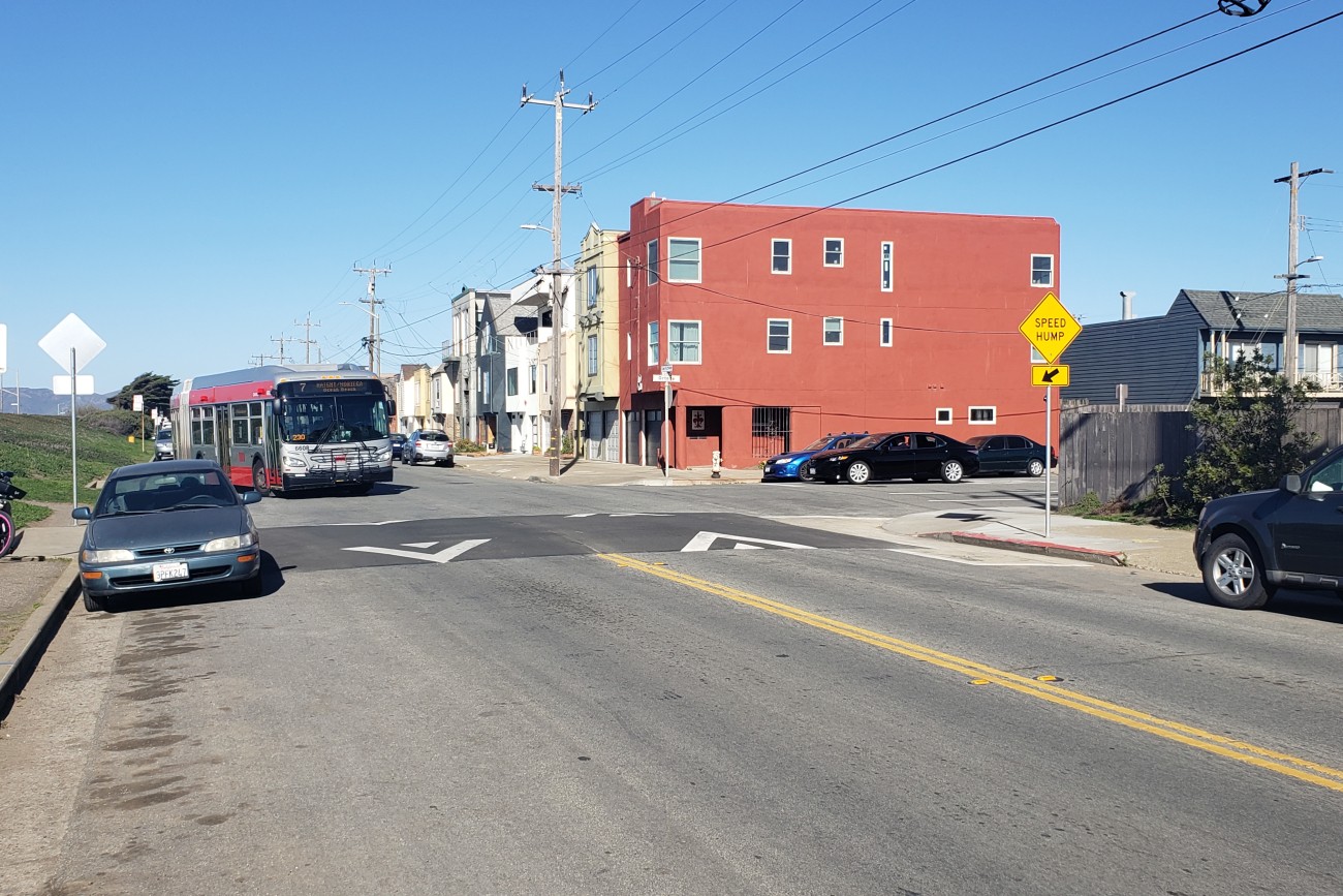 The intersection of Great Highway and Ortega on a sunny day. The 7 Muni bus approaches the intersection, which sports a new speed table crossing.