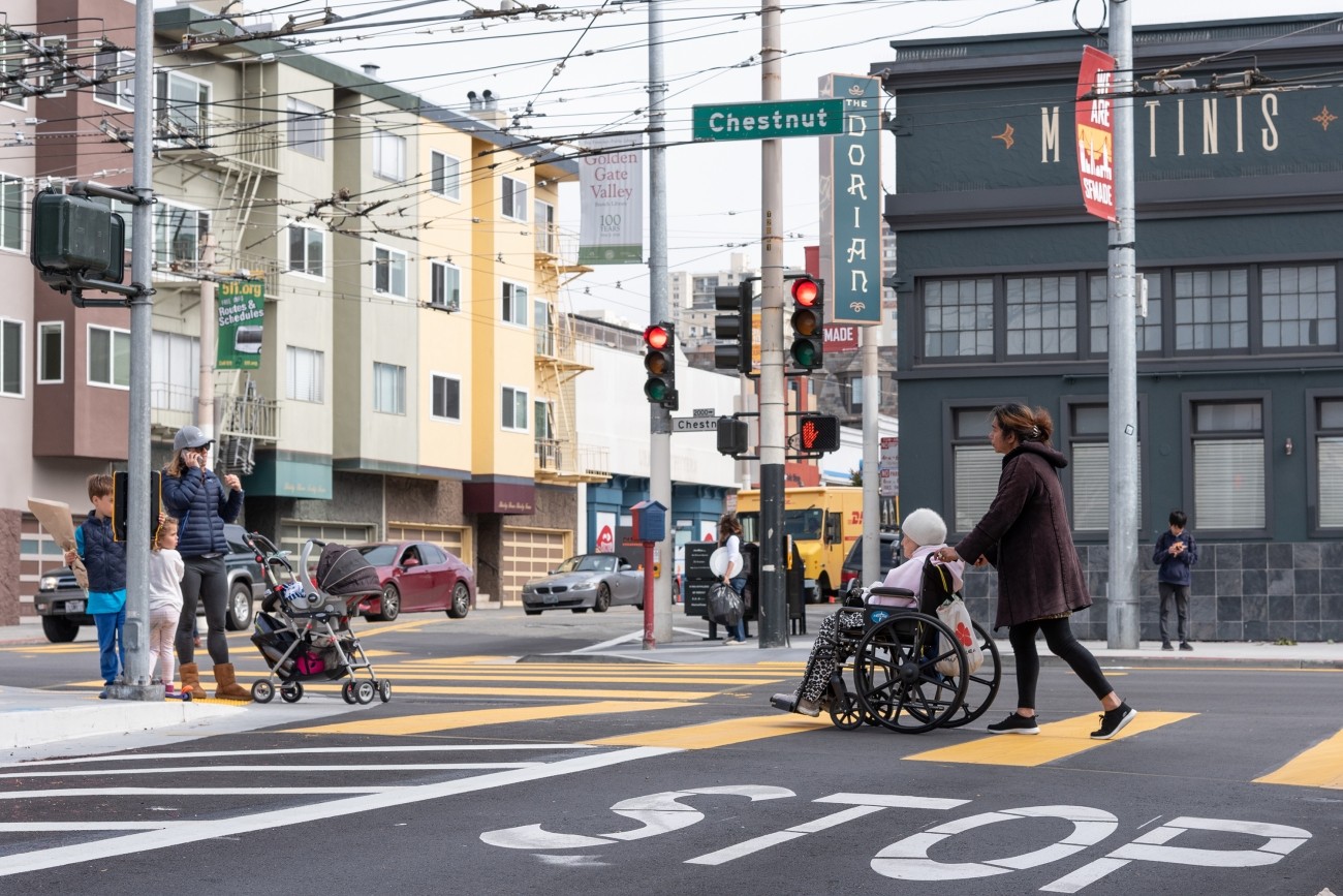 A parent with two kids and a stroller wait at the corner of Chestnut and Fillmore, while a young woman pushes an elderly woman in a wheelchair in the crosswalk.