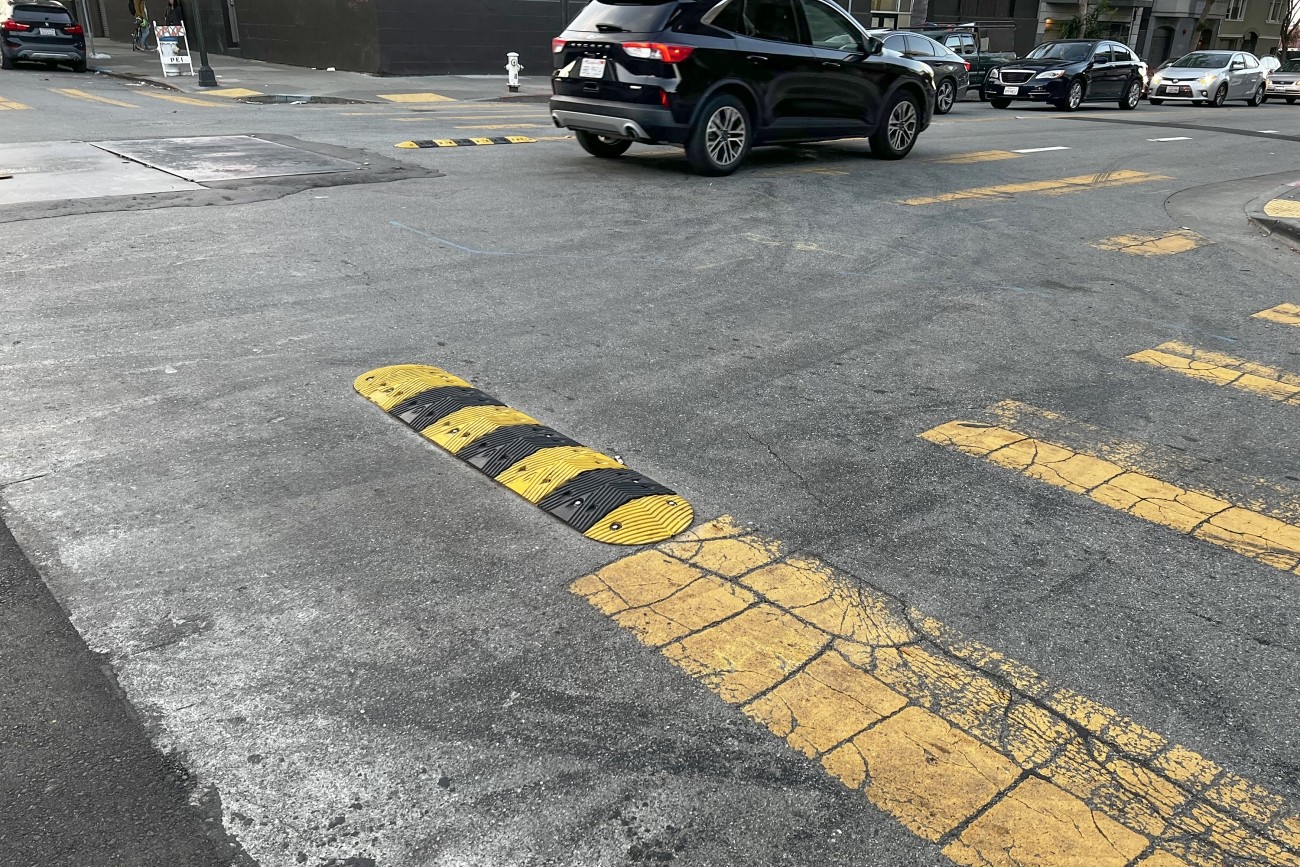 Eye-level view of an intersection, the pavement streaked with sideshow tire markings. Black and yellow striped sideshow deterrence humps, about 4 feet long, 1 foot wide, and a few inches high, protrude into the intersection from the corners and between the two lanes of each street.