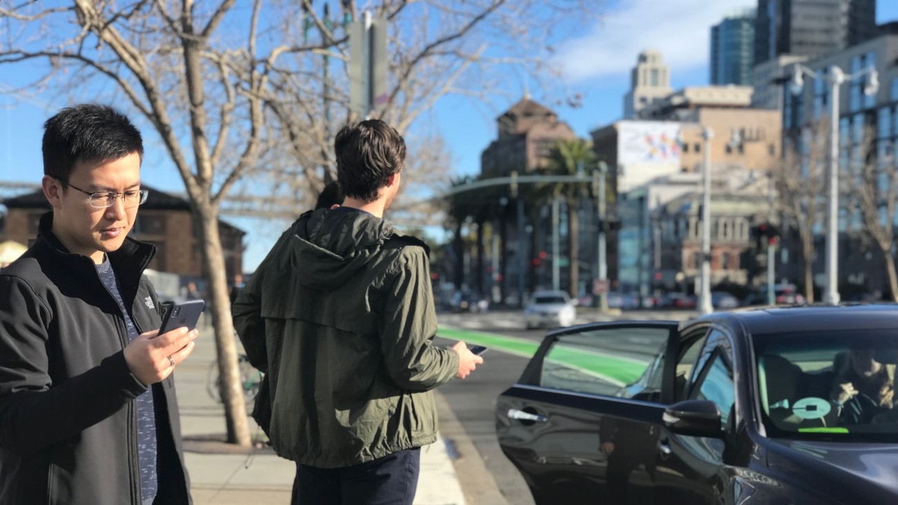 People standing on a sidewalk with phones and a ride-hail vehicle dropping someone off