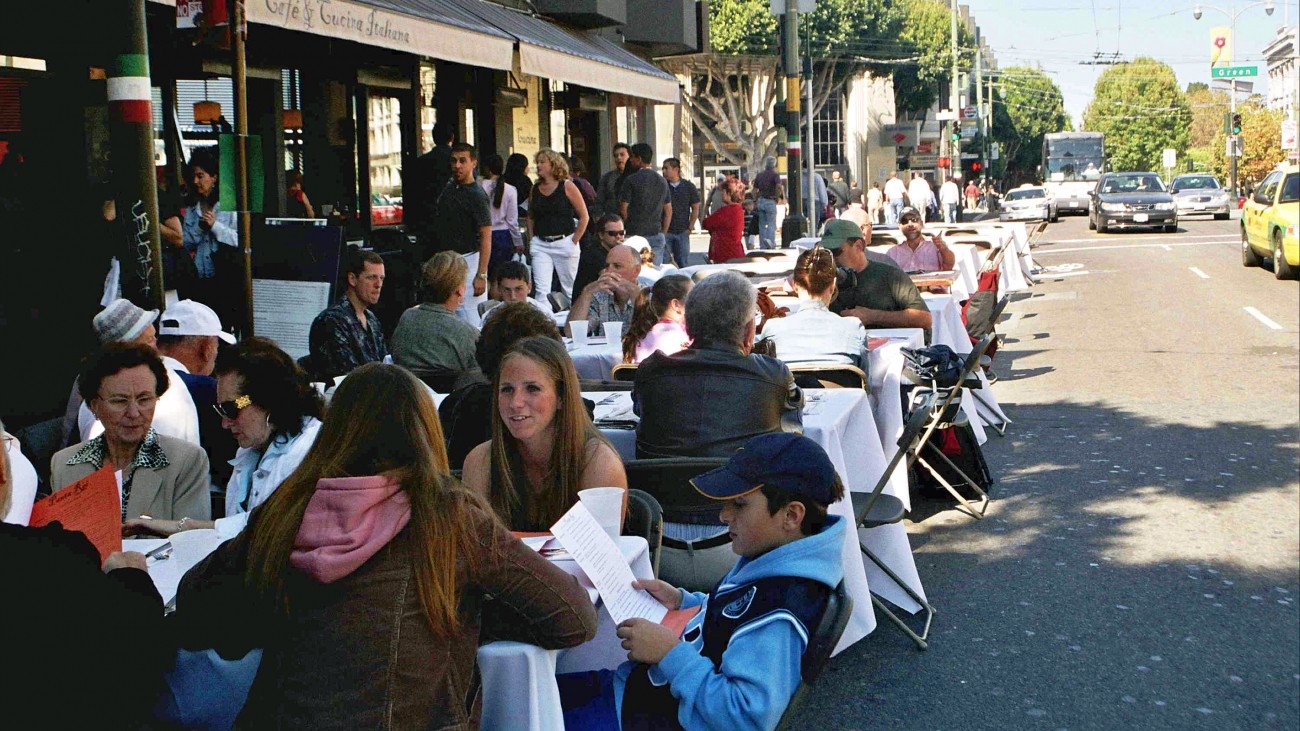 People eating on tables in parking spots in front of a cafe 