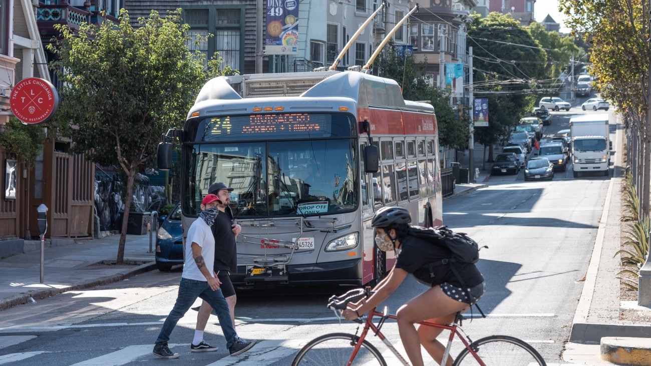 24 Divisadero Muni bus in background with pedestrians and a bicyclist in foreground