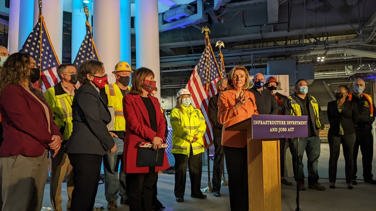 Speaker Pelosi speaking at the podium with city officials and agency staff gathered around.