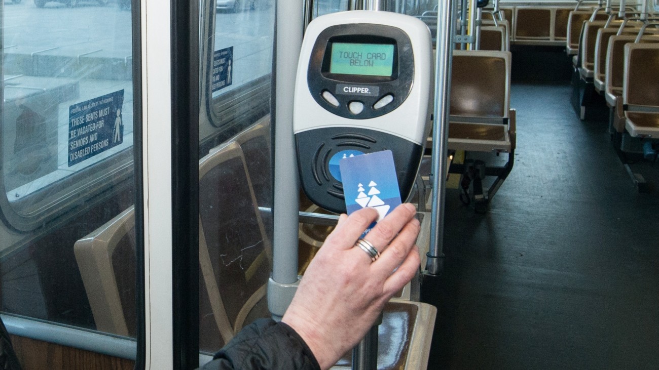A hand holds a Clipper card up to a wireless card reader. The card reader display reads "touch card below." Photo by SFMTA Photography Department