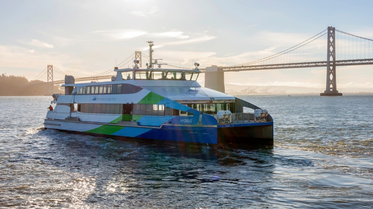 An SF Bay Ferry vessel cruises in front of the Bay Bridge. The early morning sun backlights the scene. Photo by Sergio Ruiz: flic.kr/p/2gbGJHK