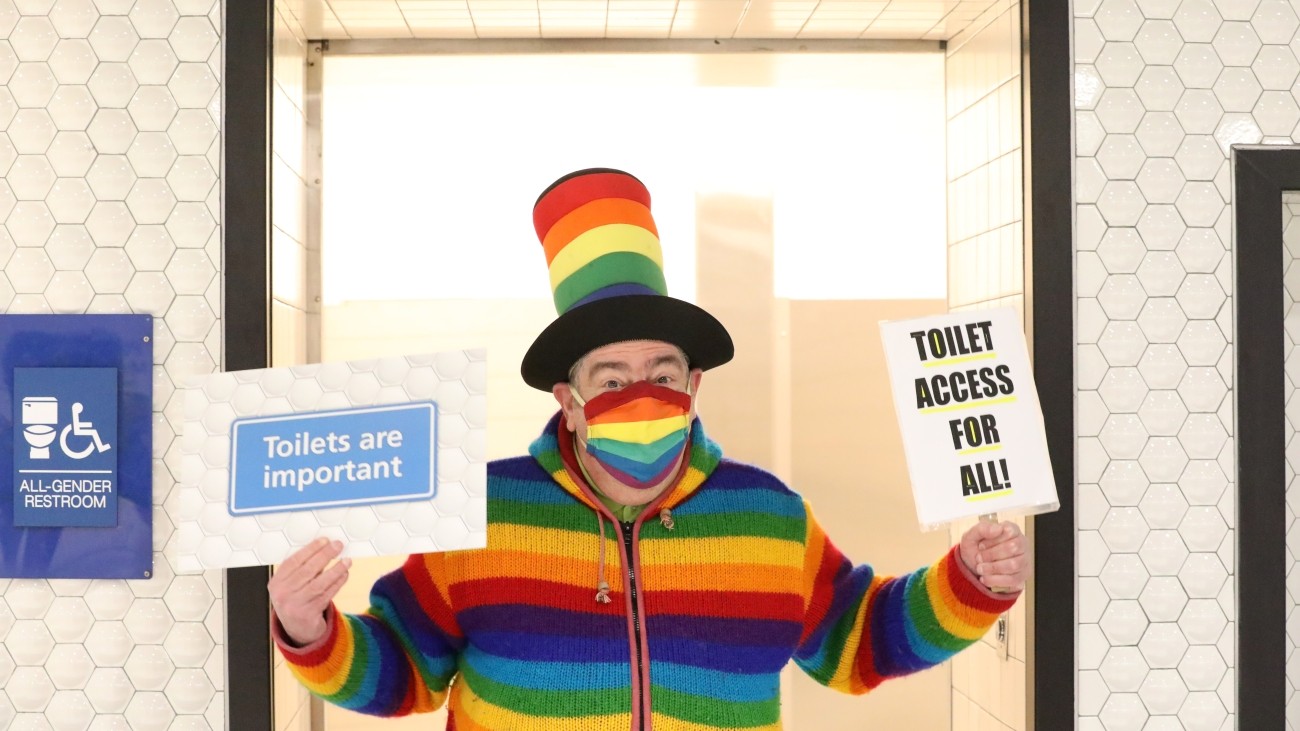Man in rainbow-colored attire holds signs in front of the restroom that says "toilet access for all" and "toilets are important."