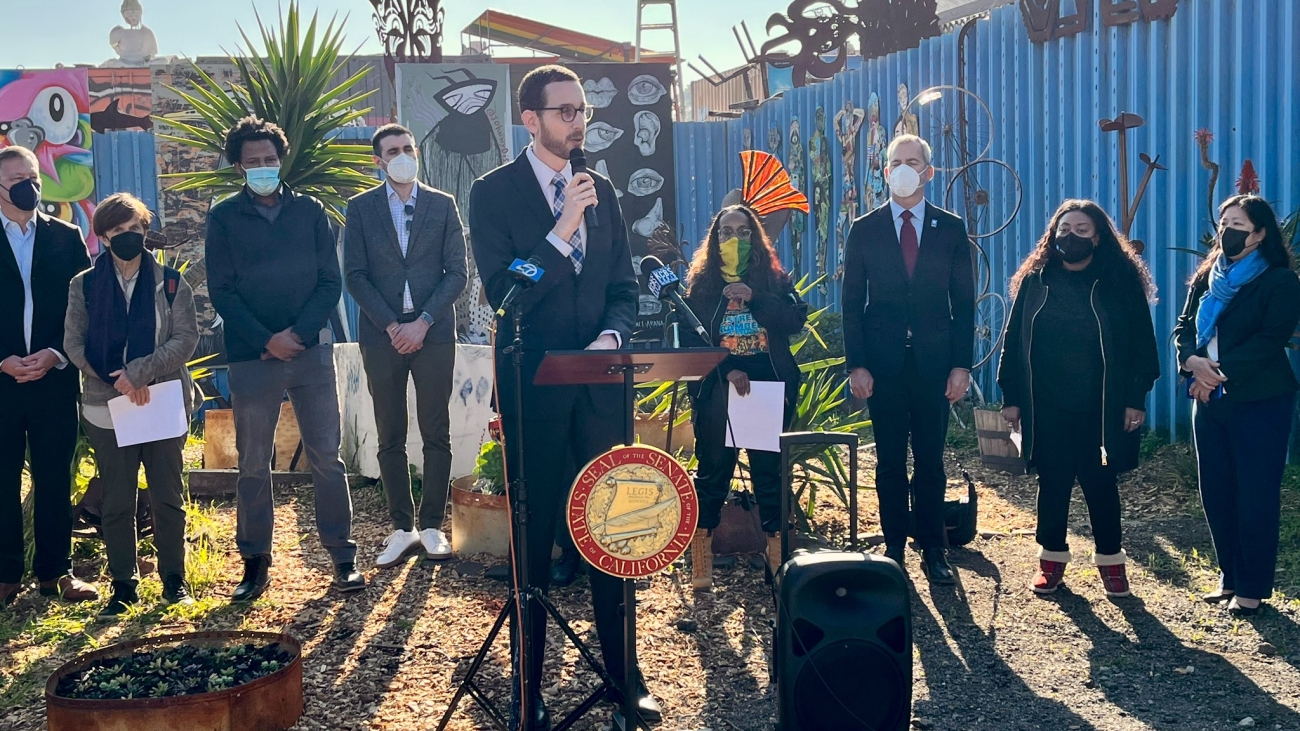 Senator Scott Wiener giving a speech in front of outdoor artworks as community leaders and transit officials gather behind him.